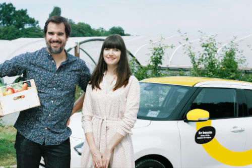 Un homme et une femme devant une Yelomobile garée devant une serre.
