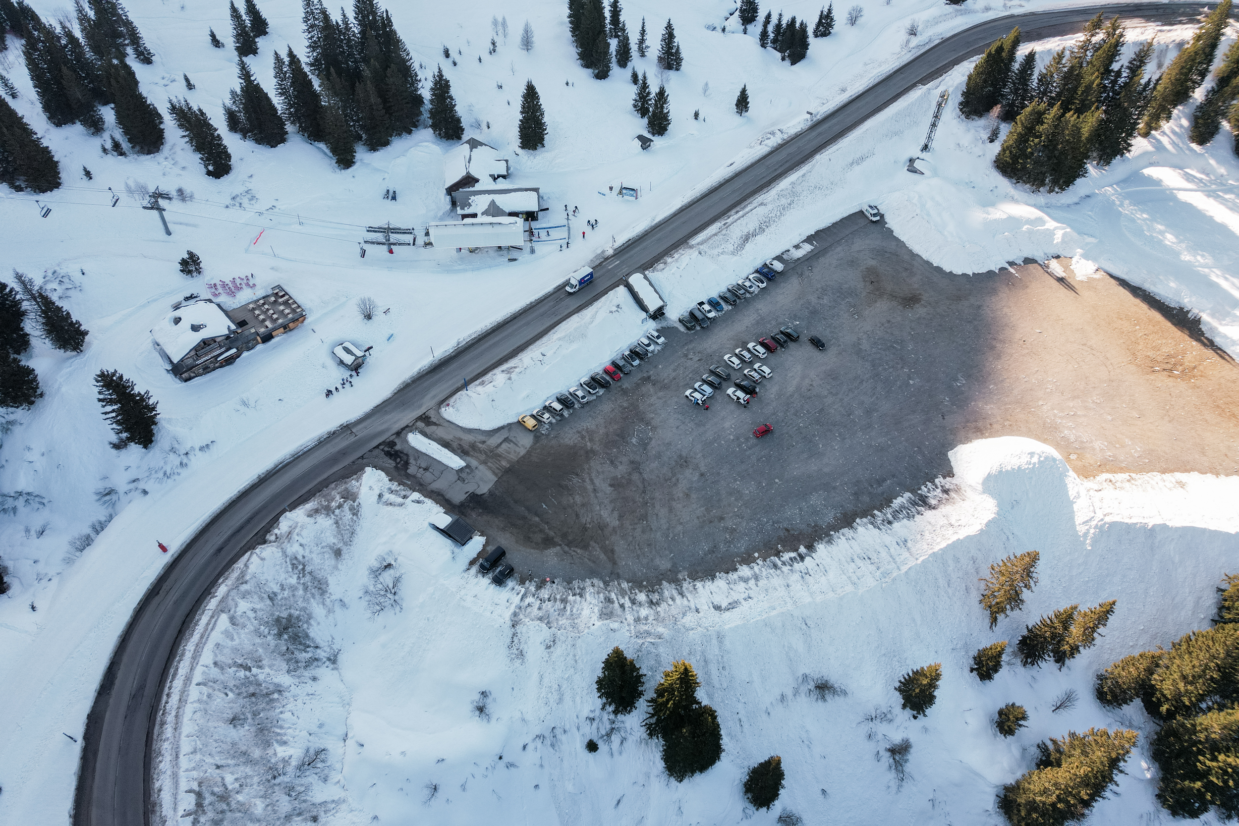 Une station de ski vue du ciel avec un parking au milieu de la neige. Une route passe à côté de quelques chalets et de remontées mécaniques.