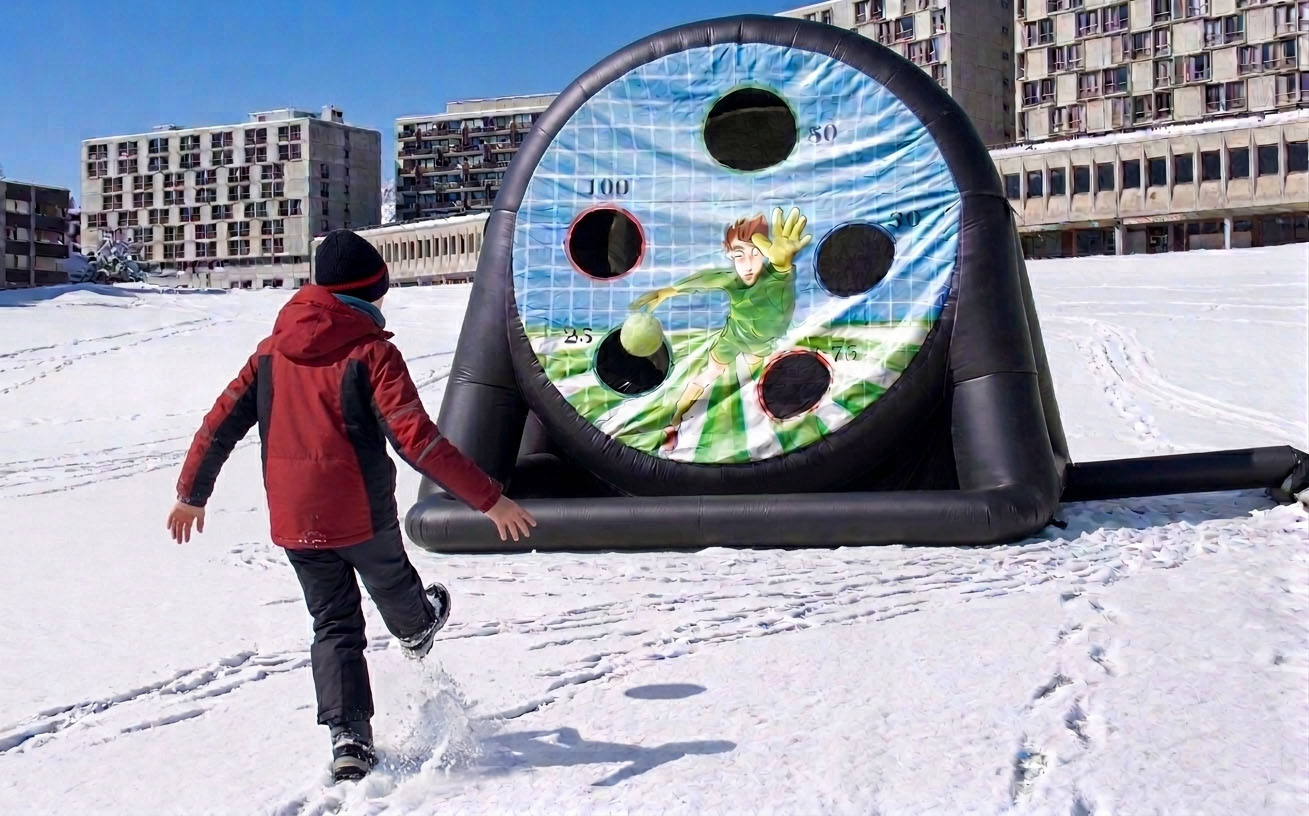 Een kind in het rood schopt in de sneeuw naar een grote opblaasbare schijf met een getekende keeper. Op de achtergrond staan gebouwen.