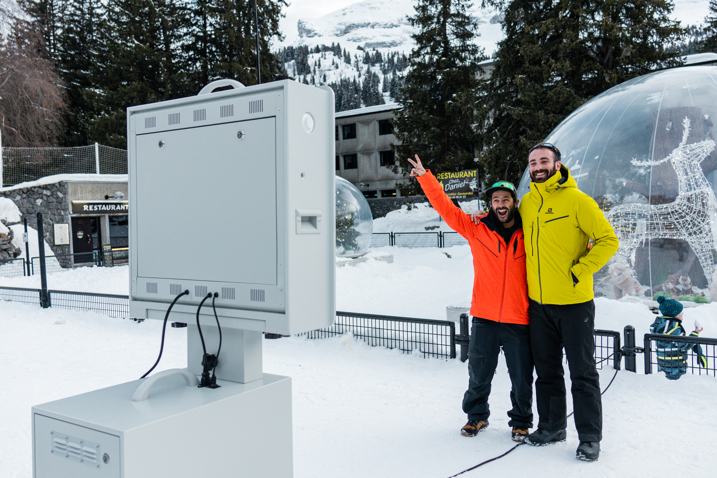 Twee mannen in ski-uitrusting glimlachen en poseren voor een fotocabine in een besneeuwd bergstation.