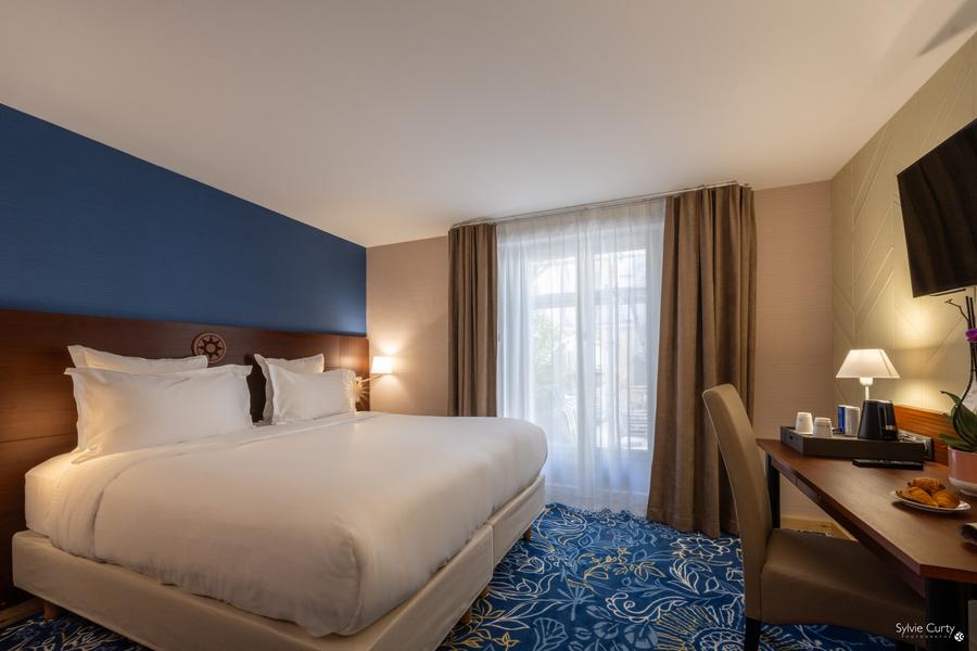 Une chambre avec un lit double, télévision, bureau, chaise, porte-fenêtre.