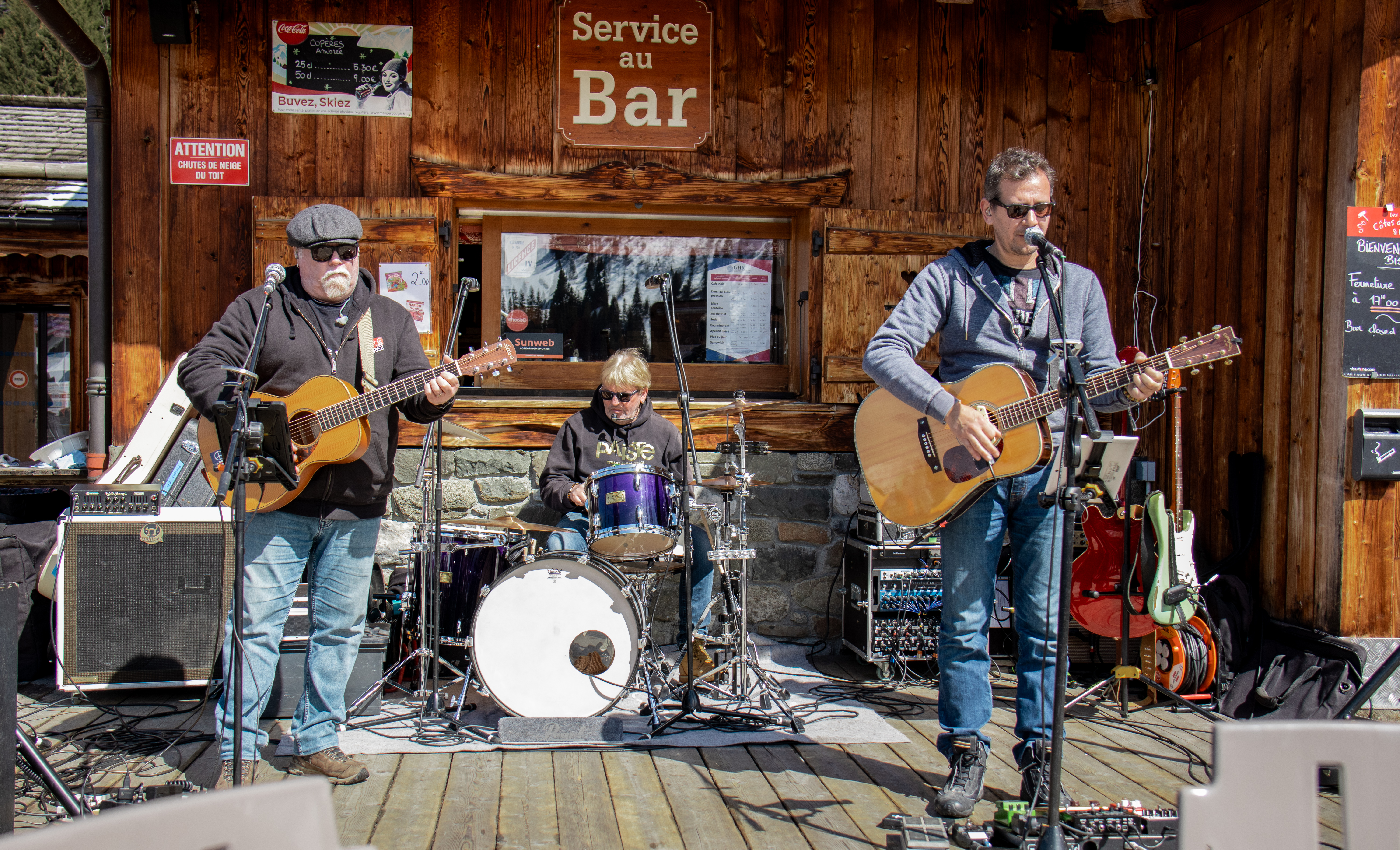 Trois musiciens jouent sur une terrasse en bois devant un bar de montagne. Le groupe se compose de deux guitaristes, chanteurs et d'un batteur au centre.