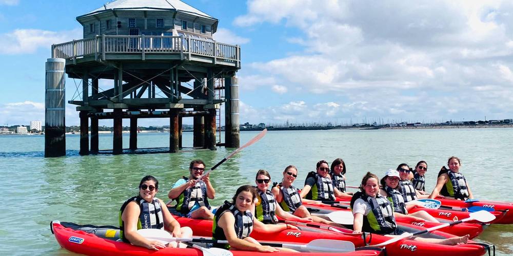 Une douzaine de personnes sur des canoes, sur l'eau devant la réplique du phare du bout du monde