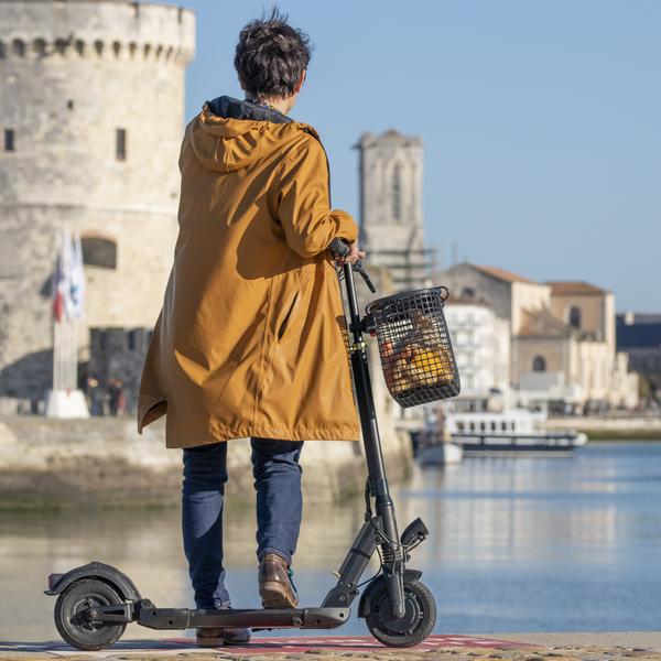 Une personne en jaune de dos face à la tour St Nicolas, avec une trotinette portant un  panier en matière de poches ostéricole.