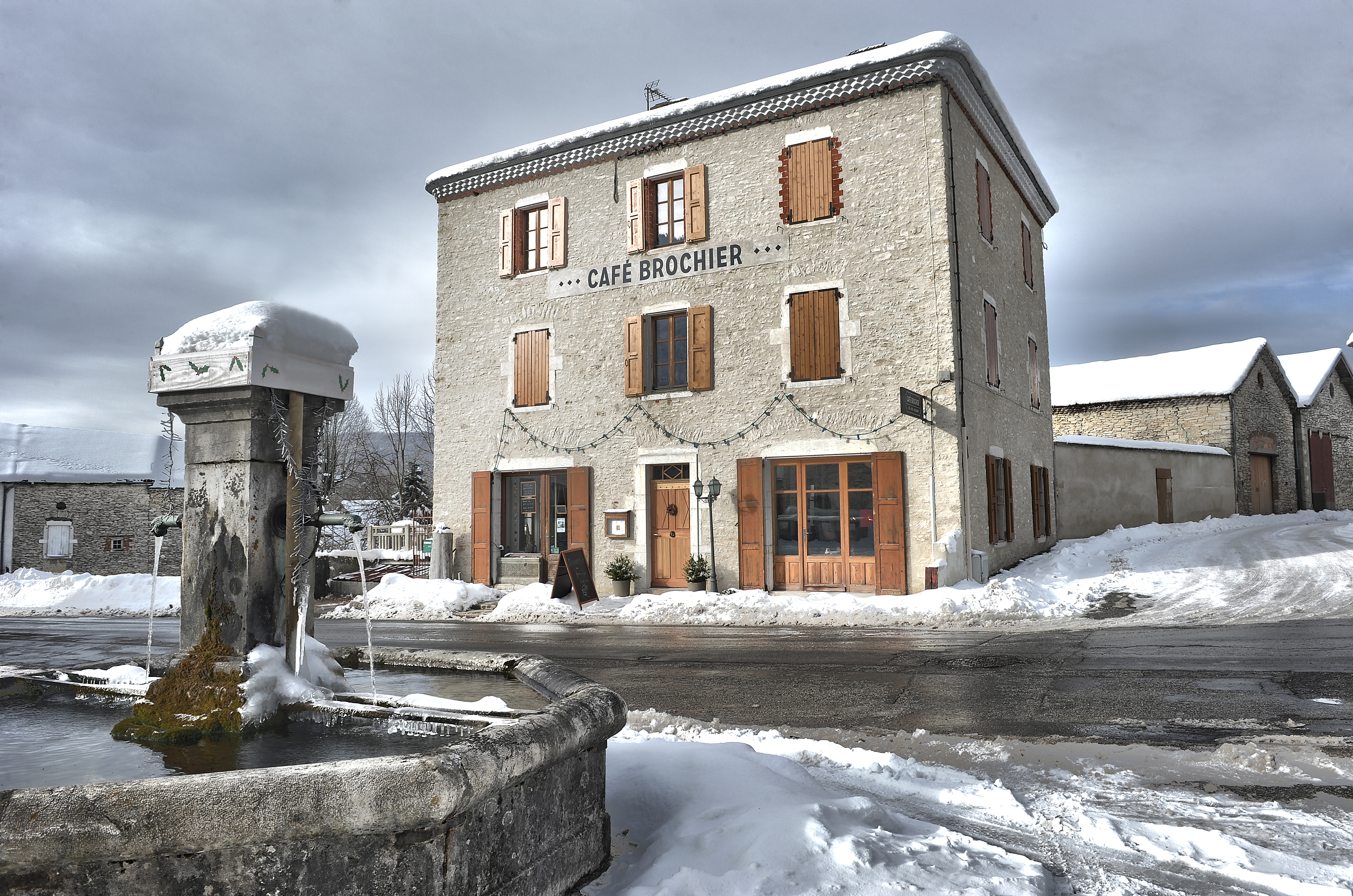 Ambiance hivernale avec une fontaine recouverte de neige au premier plan et l’hôtel restaurant visible en arrière-plan de l’autre côté de la route