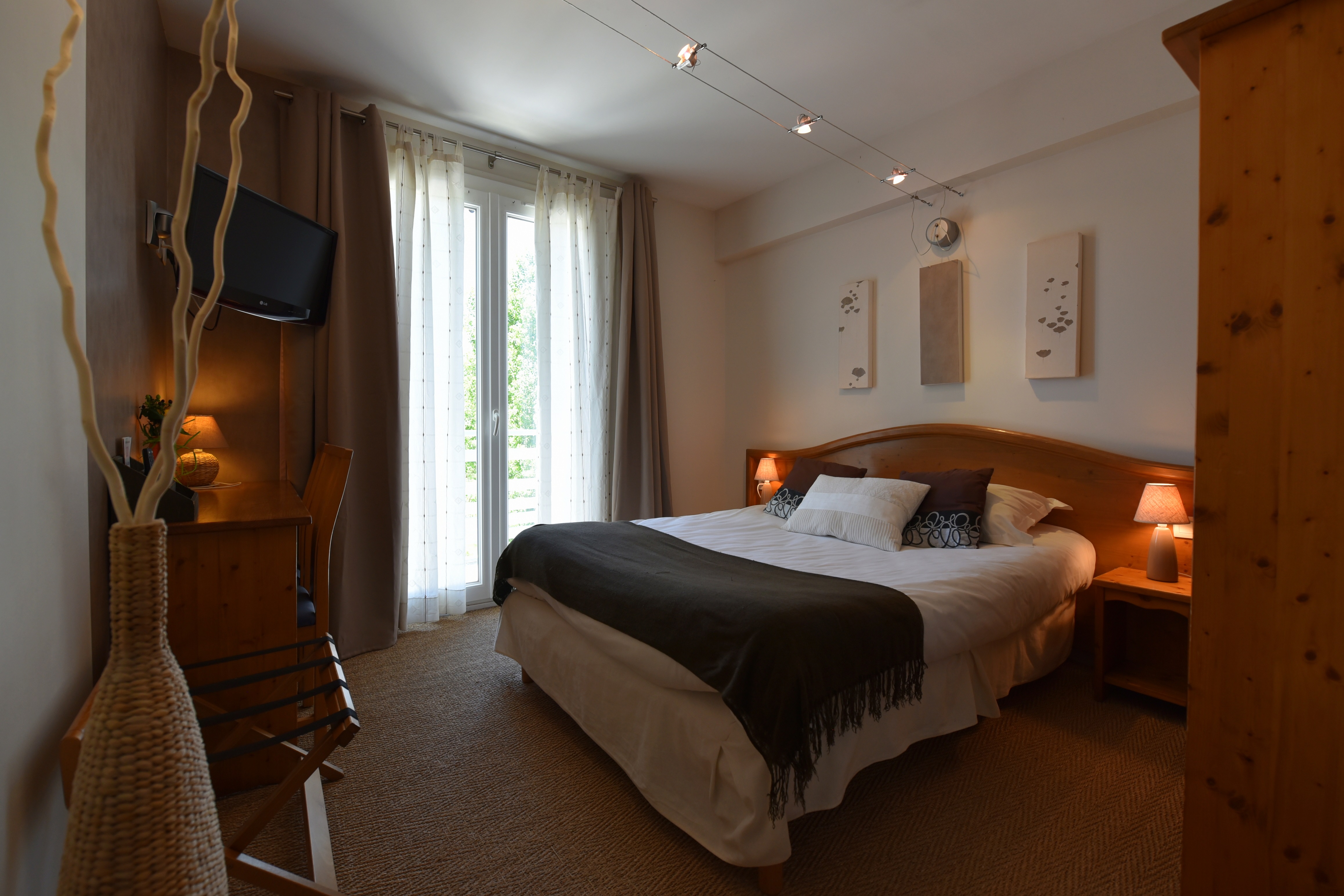 Chambre double chaleureuse équipée de deux lampes de chevet et d’une lampe de bureau allumées, de trois cadres décoratifs au-dessus du lit, d’une armoire, d’un porte bagage et d’un téléviseur. Trois spots orientables éclairés sur rail assurent un éclairage central. Vue sur la verdure par la porte-fenêtre