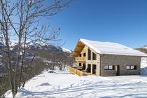 Vue de côté d'un grand chalet en bois et pierre sur deux étages, nombreuses fenêtres et large terrasse, balcon au 1er étage.