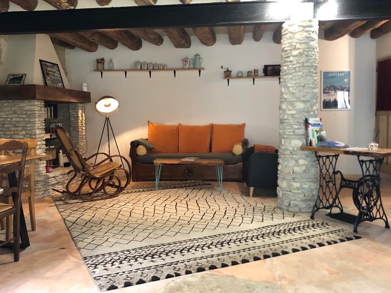 Salon chaleureux avec mobilier en bois, tapis géométrique, fauteuils, plantes et décoration murale évoquant une ambiance artisanale et conviviale.