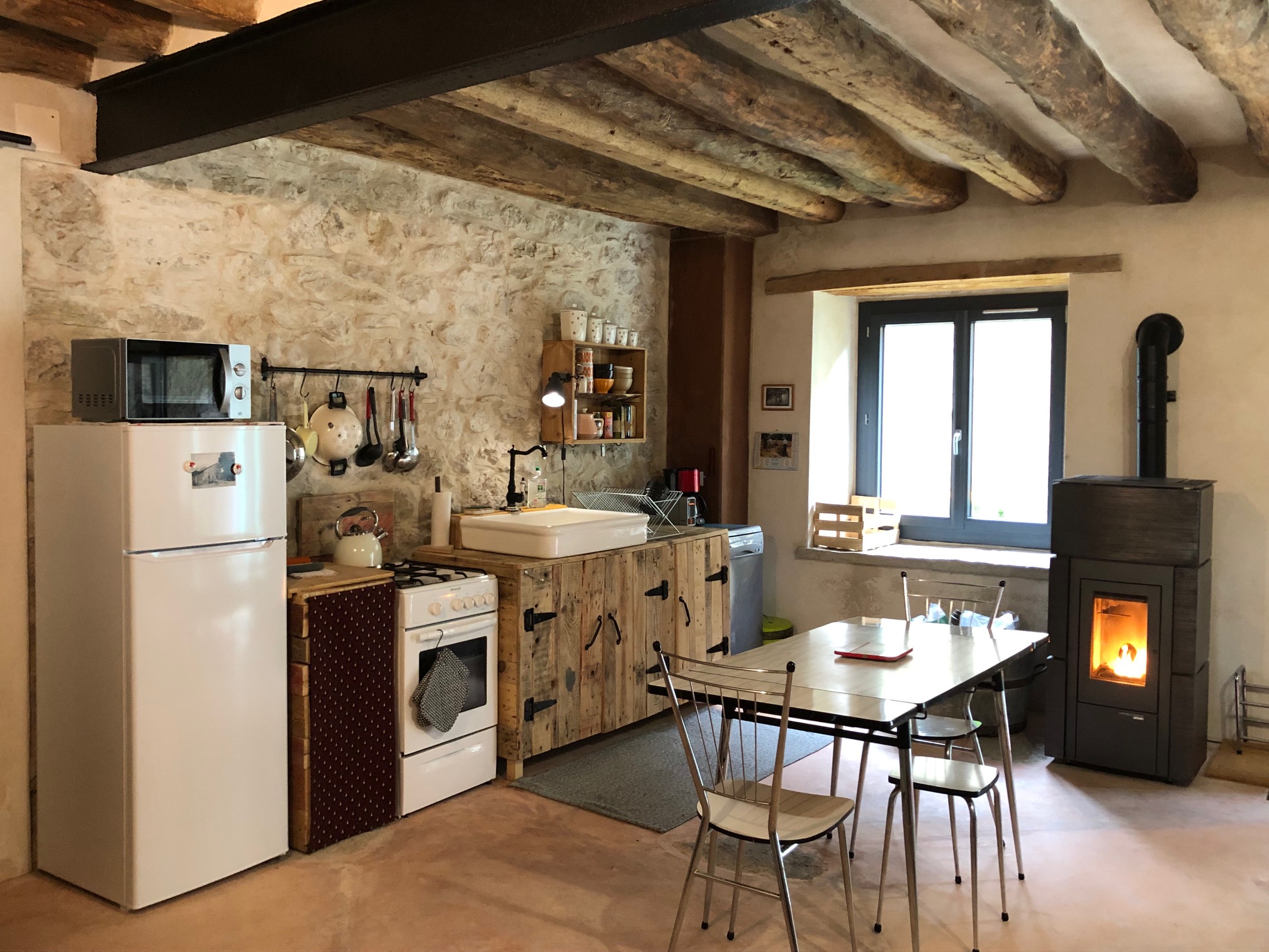 Cuisine rustique et lumineuse avec poutres apparentes, meubles en bois, réfrigérateur, table conviviale, grandes fenêtres et poêle chaleureux.