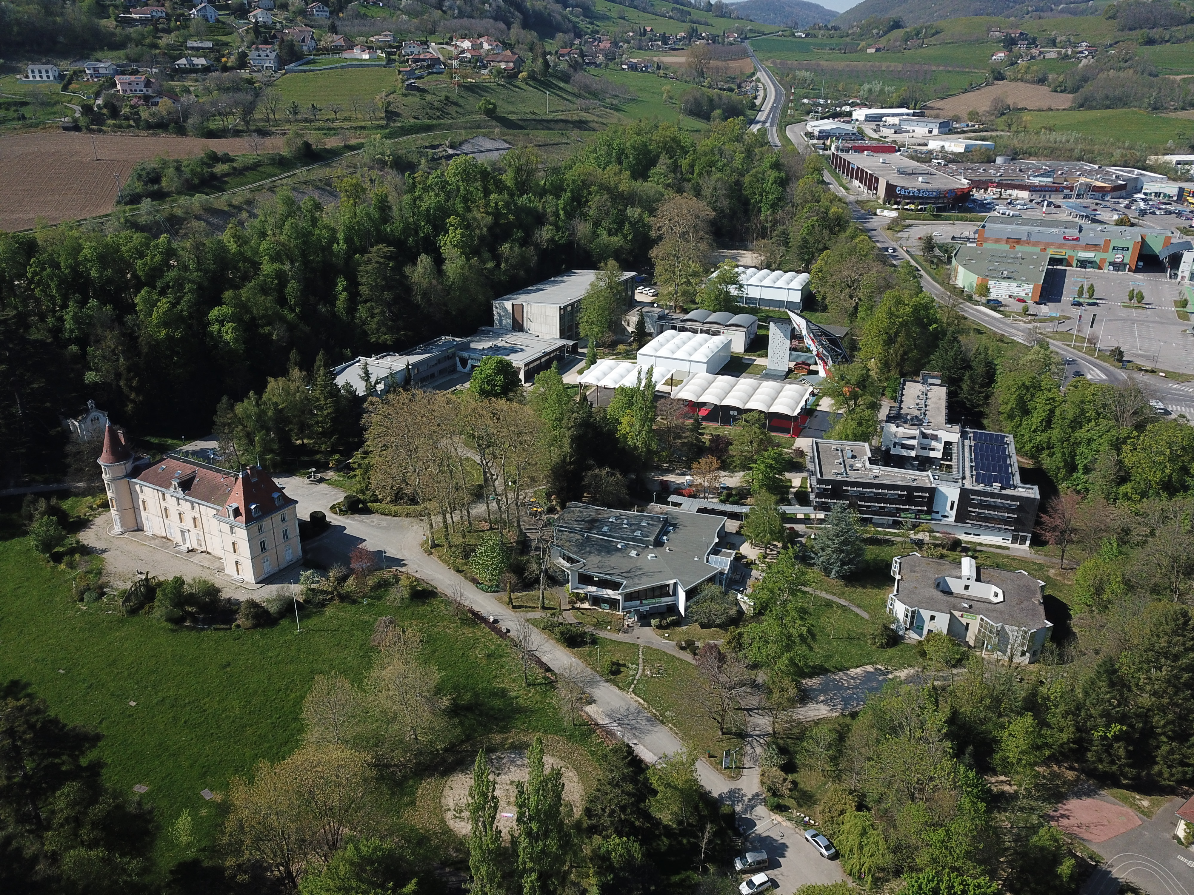 Une vue aérienne présente un grand campus aux multiples complexes, entouré d'arbres et bordé par une grande route reliée à un centre commercial