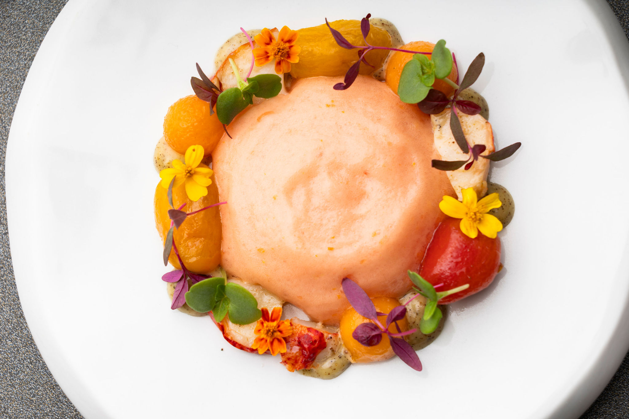 Au centre d'une assiette blanche, un cercle de mousse légère de couleur pêche avec petites feuilles et fleurs comestibles de couleurs vives.