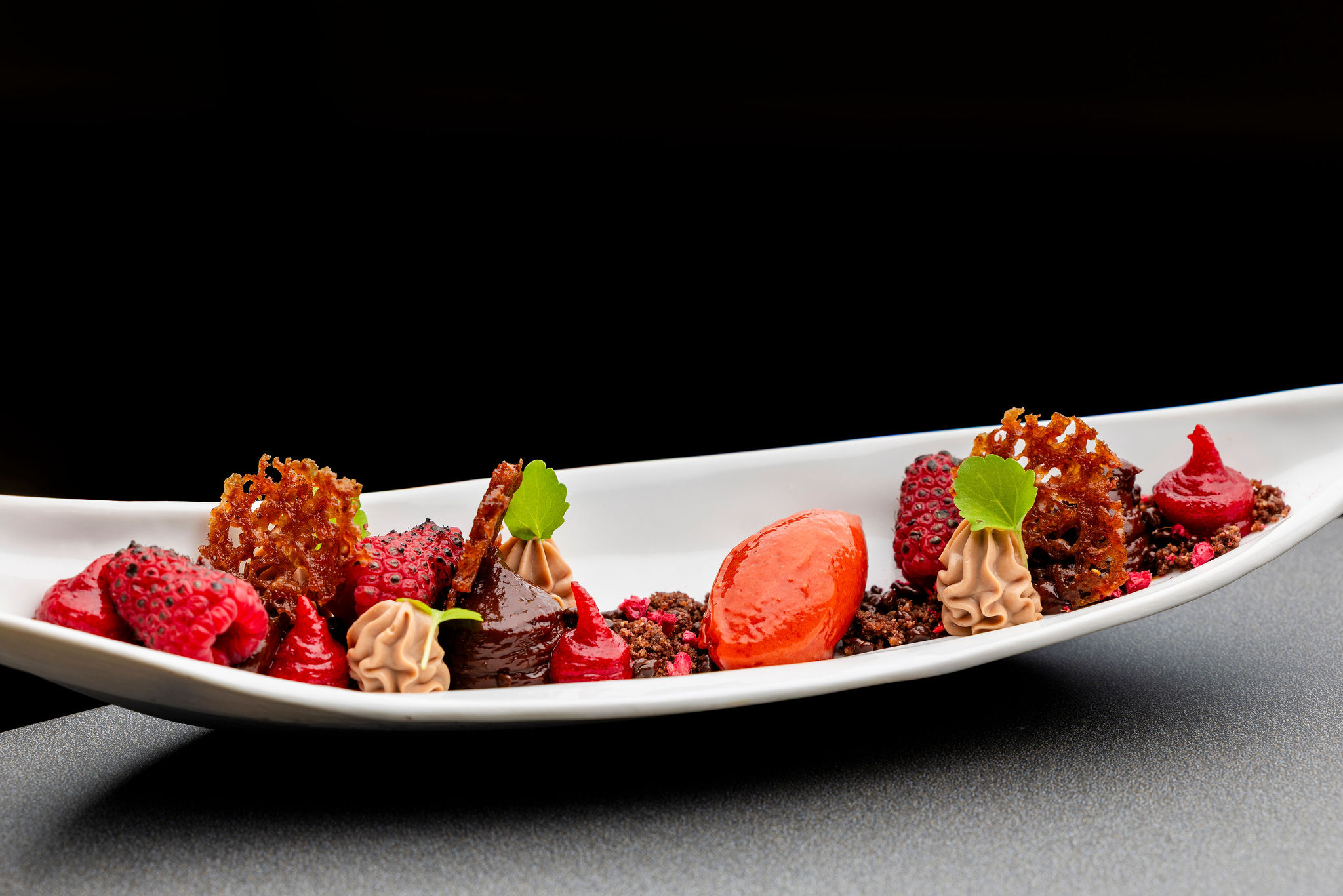 Un dessert artistique est présenté sur une longue assiette blanche incurvée, composé de plusieurs éléments dont une quenelle rouge vif, des framboises entières, des pointes de crème légère, des morceaux de tuile croustillante et des miettes foncées.