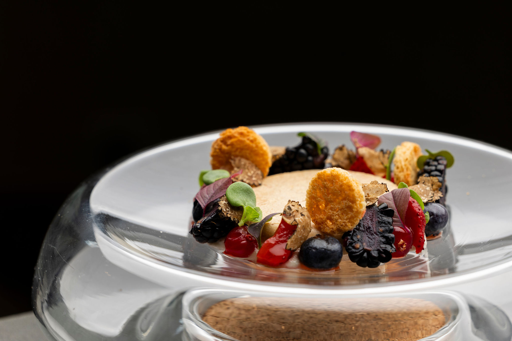 Sur un socle circulaire transparent, une petite assiette en verre contient un dessert en forme de couronne, composé d'une base claire recouverte d'un anneau de fruits des bois noirs et rouges, de petites tuiles rondes dorées et de quelques feuilles vertes.