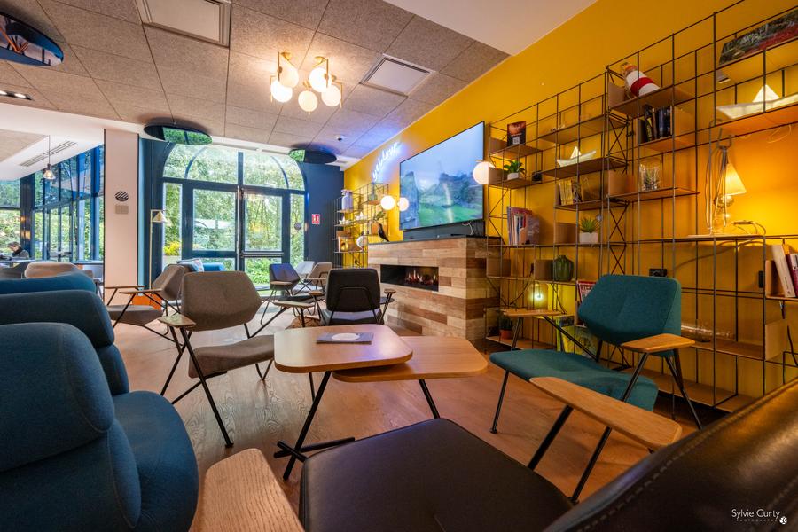 Un coin repos dans un espace commun de l'hôtel, avec des tables basses et fauteuils, un grand écran de télévision.