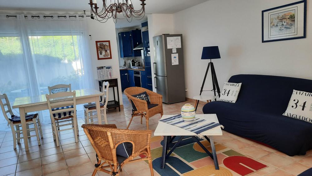 Salon - séjour avec canapé, deux sièges en osier, une table basse, une table à manger avec quatre chaises, au fond la cuisine.