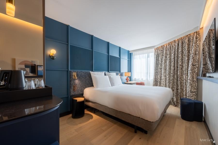 Une chambre avec un lit double. Un mur est bleu, un grand rideau ferme une partie de la grande fenêtre.