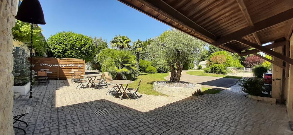 Le jardin vue depuis le préau, avec des tables et des chaises en bois, sur la terrasse, et un espace arboré.