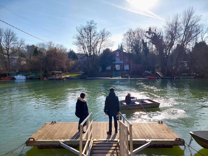 Deux personnes attendent sur un ponton flottant de monter à bord d'une barque pour traversée la Marne vers l'atelier
