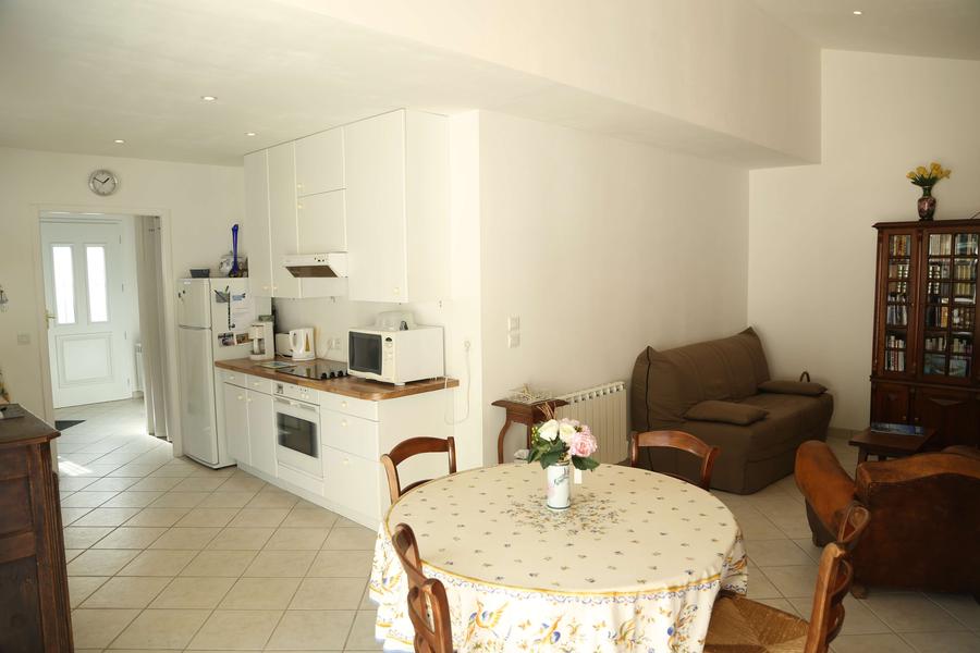Pièce de vie avec cuisine ouverte tout équipée, table ronde avec 4 chaises, canapé, fauteuil, bibliothèque.