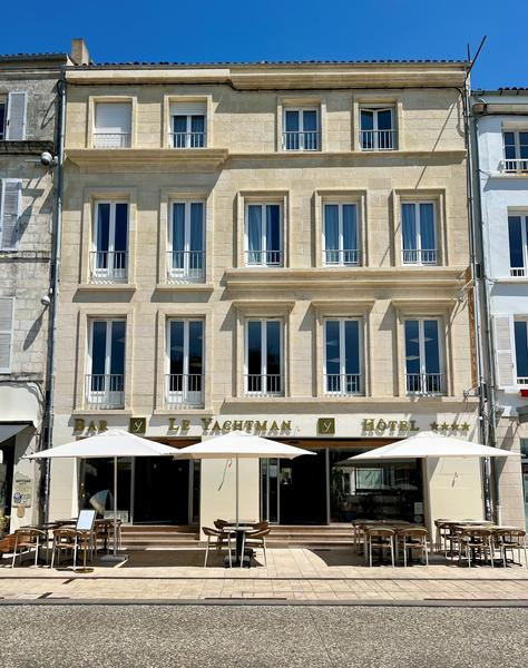 La facade de l'hôtel depuis la rue, avec des tables, chaises et parasols sur la terrasse.