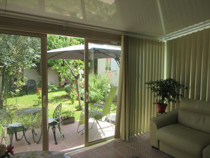 Autre vue de la véranda, avec à l'extérieur des transats, une table et deux chaises., un parasol. Le jardin est fleuri et avec des arbres.