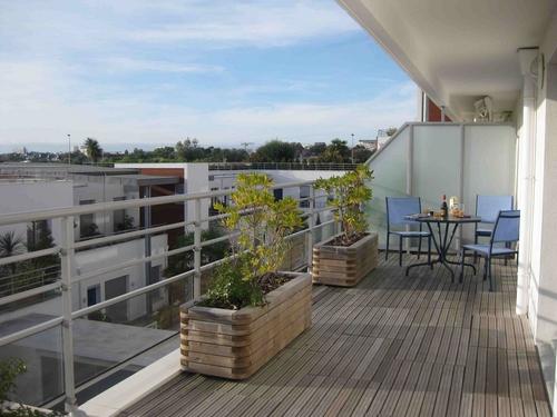Le grand balcon avec caillebotis en bois, grandes plantes, et  au fond une table et trois chaises.