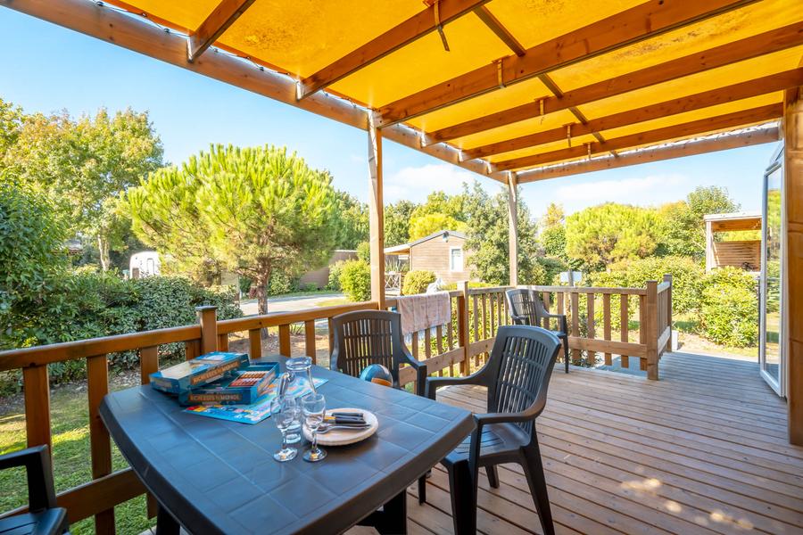 la terrasse couverte d'un mobil home, avec une table d'extérieur: des verres et des jeux de société sont dessus.