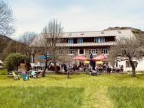 Vue du restaurant en été, grande bâtisse sur 3 étages en pierre, terrasse extérieure avec tables et chaises, en fond de prairie