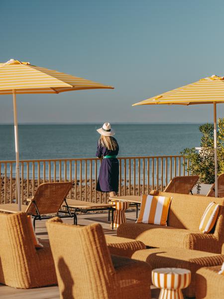 Une dame de dos appuyé contre la rembarde, sur la terrasse, avec des transats, parasols, tables et chaises.