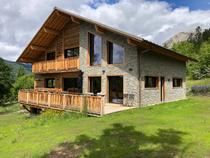 Grand chalet en bois et pierre sur deux étages, nombreuses fenêtres et large terrasse, balcon au 1er étage. Paysage printanier.