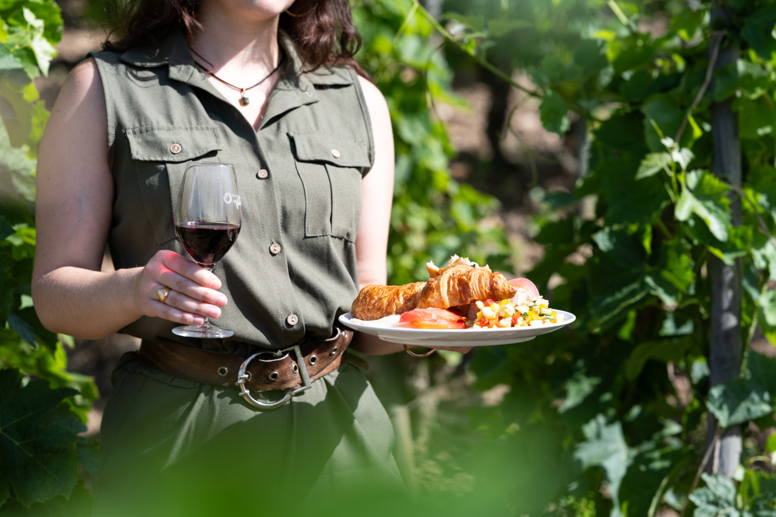 Une femme, dans les vignes, tient dans une main une assiette blanche remplie de nourriture et dans l'autre main un verre de vin rouge