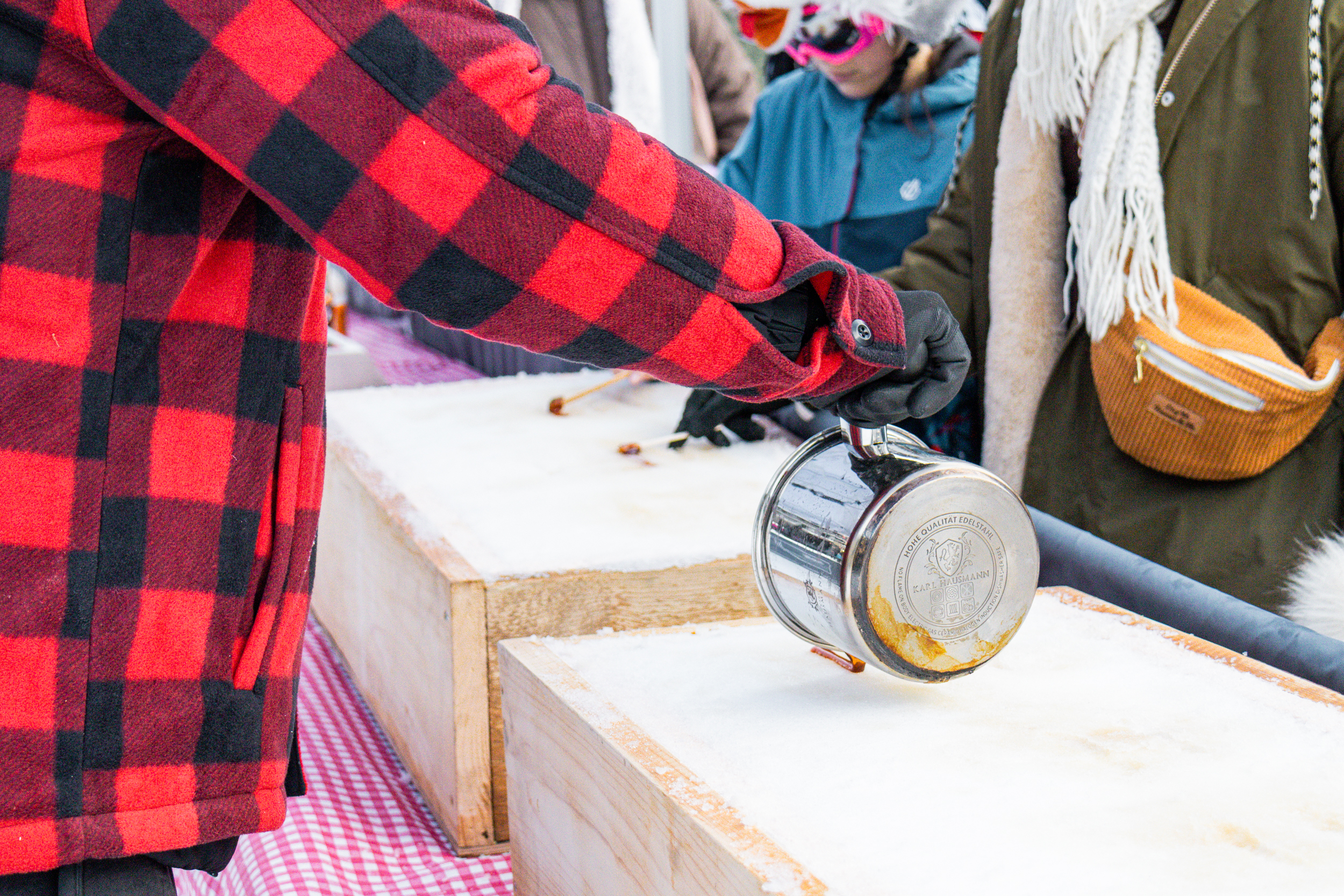Iemand met een rood-zwart geblokte jas giet warme siroop op verse sneeuw in houten bakken om 