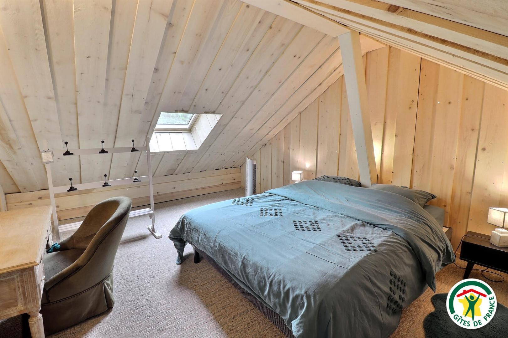 La chambre mansardée de l'étage : lit 140x190cm, velux avec store, fenêtre avec rideaux, portant, bureau