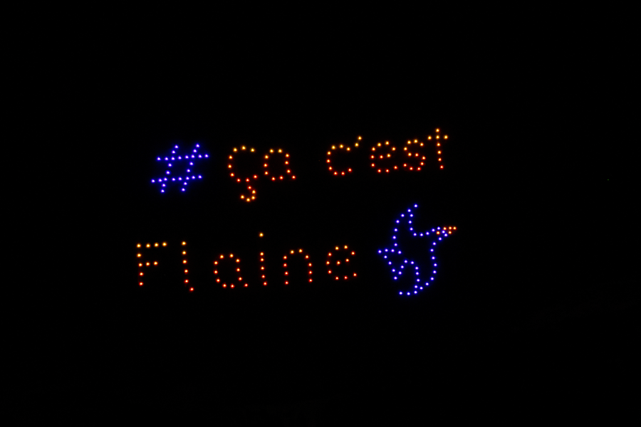 Spectacle de drones lumineux dans un ciel nocturne totalement noir. Les points de lumière forment le texte en français « # ça c'est Flaine » écrit en orange et bleu.