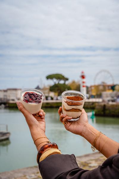 Une personne tenant un tiramisu dans une main, et un pana cotta dans une autre, face au port.