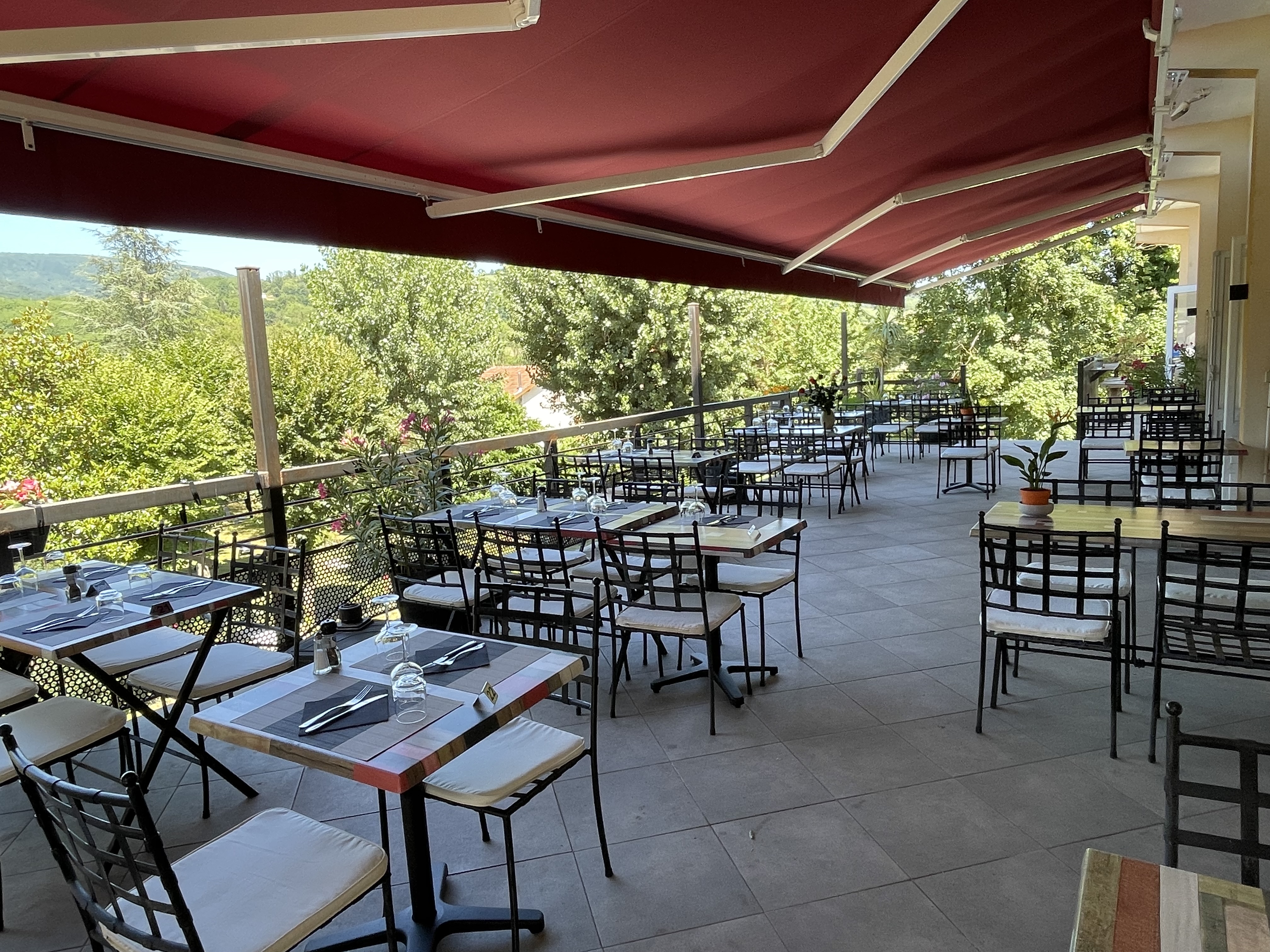 Terrasse conviviale, protégée du soleil par un large store, où une dizaine de tables sont installées, offrant une vue sur le parc arboré