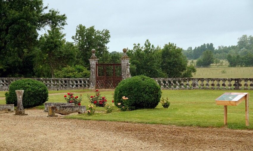 Esplanade de verdure entourée dun muret en pierre, arbustes, fleurs et bancs de pierre sur le devant, grands arbres au fond