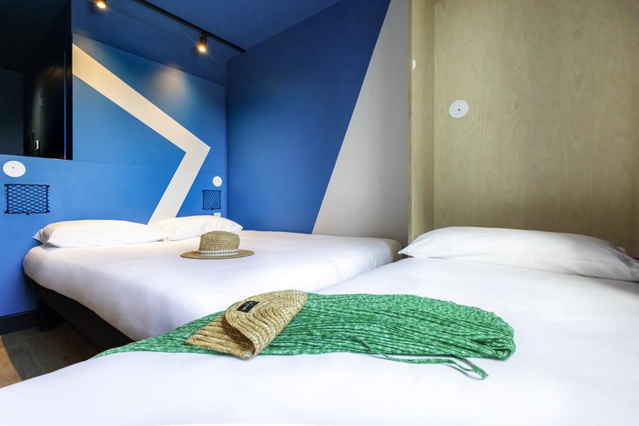 Une chambre avec un lit double et un lit simple. Un paréo et un sac sont posés sur le lit simple, un chapeau est posé sur le lit double.