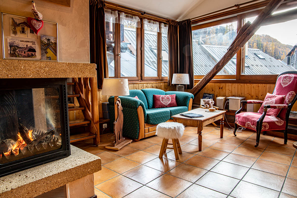 Séjour chaleureux et lumineux avec canapé, fauteuils, cheminée, fenêtres donnant sur le village