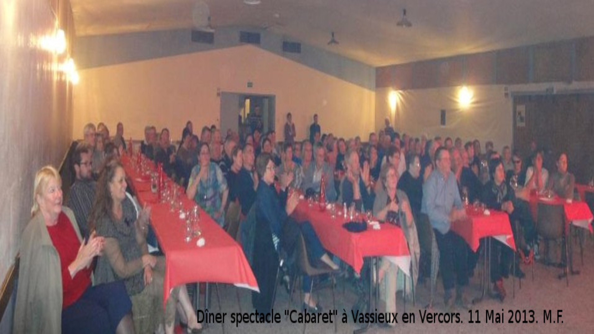 Des adultes sont assis dans une salle de fêtes autour de 4 grandes tables avec des nappes rouge, tous regardent dans la même direction un spectacle.