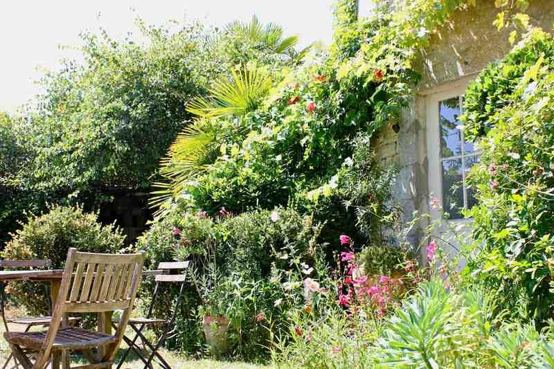 Une partie d'un jardin arboré, avec un salon de jardin en bois, la végétation grimpant sur la façade du moulin.