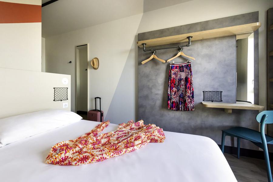 Une chambre avec un lit double, une robe est posée dessus, un bureau, une chaises et quelques cintres, une jupe accrochée à l'un.