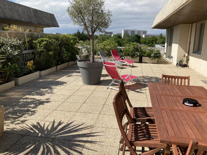Très grand balcon avec arbustres, un arbre en pot, trois bains de soleil, et une table avec 6 chaises en bois.