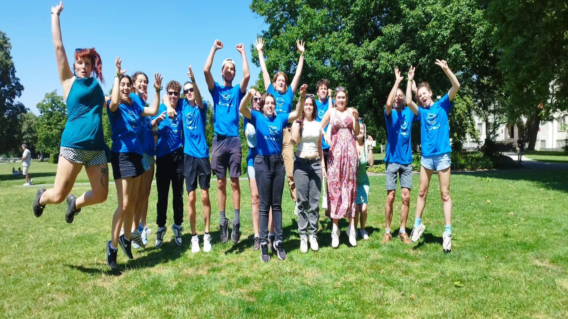 Dans un parc, un groupe de jeunes de l'association Activ'Royans tous portant des t-shirt bleus avec le logo de l'association semblent très heureux. Ils sautent et et levent les bras en l'air pour exprimer leur joie.