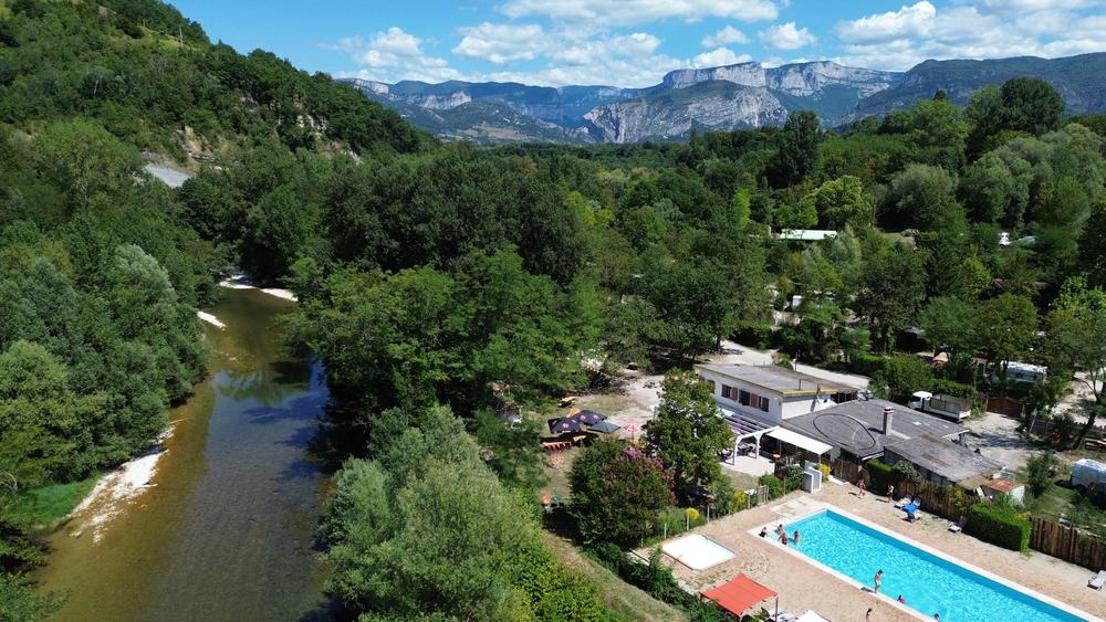 Vue aérienne du camping installé au bord d'une rivière, avec sa piscine, ses espaces arborés et les reliefs du Vercors en arrière plan