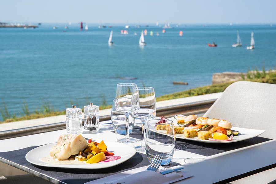 Une table face à l'océan, deux assiettes à base de poisson sont servies.