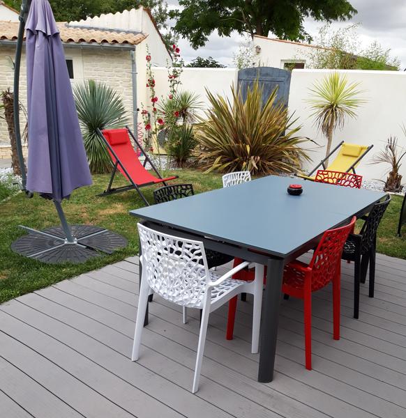 Salon de jardin sur la terrasse en bois avec table et six chaises, parasol, chiliennes, arbustes.