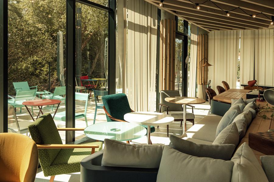 Une partie commune de l'hôtel avec des canapés, des fauteuils et des tables, avec vue sur la terrasse, ses chaises et tables métalliques.