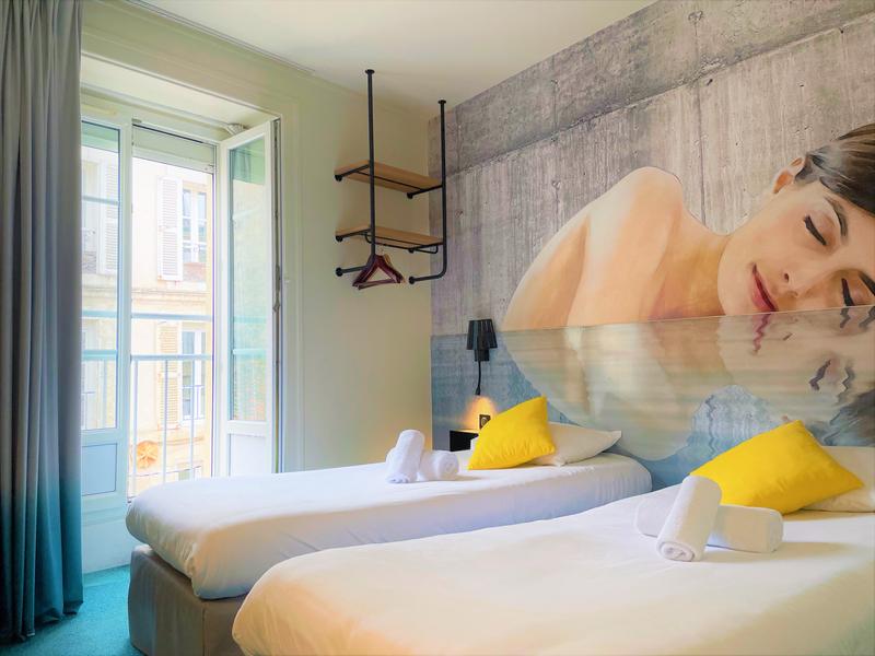 Une chambre avec deux lits simples, la porte fenêtre vers un petit balcon est ouverte. Une peinture du visage et haut du corps d'une femme au mur.