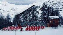 Vue du restaurant de face, chalet et terrasse en bois au milieu des pistes et de la forêt, tables et chaises, chaises longues. Montagnes en arrière-plan
